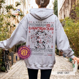 Op de afbeelding: Sportgrijze hoodie met een afbeelding van Medusa en de tekst "A Well-Read Woman is a Dangerous Creature". Het ontwerp bevat rode en zwarte accenten. De hoodie is een casual kledingstuk.