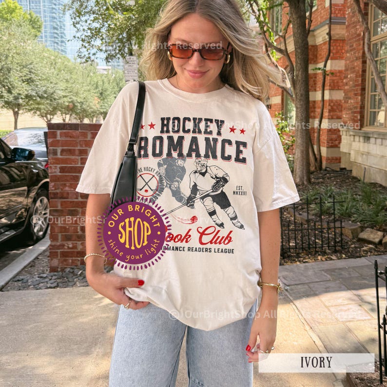 May include: Ivory-colored t-shirt with the text "HOCKEY ROMANCE" and a hockey player graphic. The shirt also features the text "Book Club Romance Readers League". The shirt is a casual, unisex style.