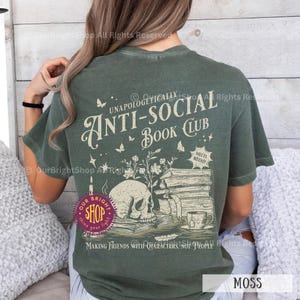 May include: Moss green t-shirt with a graphic design featuring a skull, books, and the text "UNAPOLOGETICALLY ANTI-SOCIAL BOOK CLUB." The design includes butterflies, stars, and the phrase "MAKING FRIENDS WITH CHARACTERS, NOT PEOPLE."