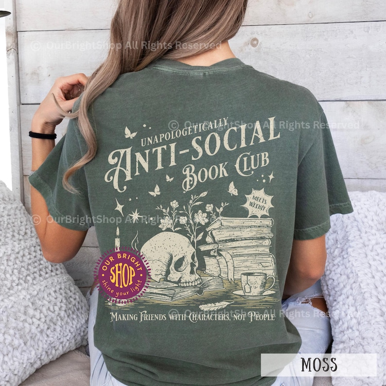 May include: Moss green t-shirt with a graphic design featuring a skull, books, and the text "UNAPOLOGETICALLY ANTI-SOCIAL BOOK CLUB." The design includes butterflies, stars, and the phrase "MAKING FRIENDS WITH CHARACTERS, NOT PEOPLE."