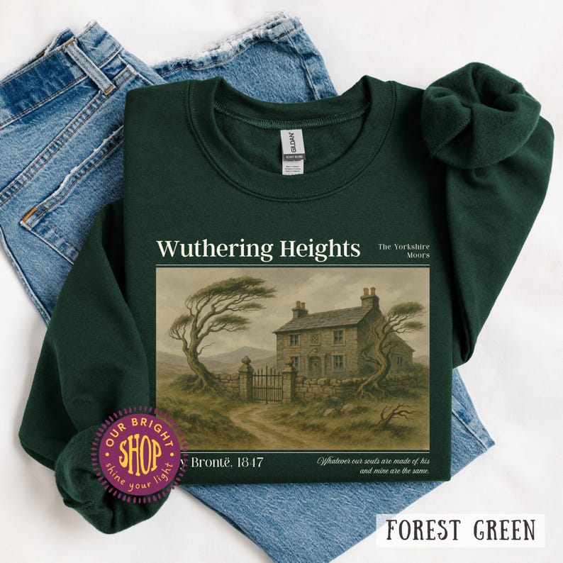 Puede incluir: Sudadera verde bosque con el t&iacute;tulo "Wuthering Heights" y una ilustraci&oacute;n de una casa y un paisaje. Tambi&eacute;n se ve el texto "Bront&euml;, 1847". La sudadera se muestra sobre unos vaqueros azules.