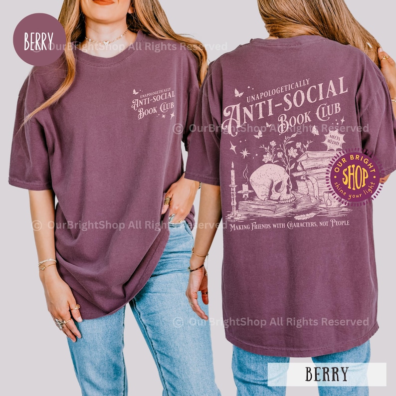 May include: Berry-colored t-shirt with short sleeves. The front features the text "UNAPOLOGETICALLY ANTI-SOCIAL BOOK CLUB". The back displays a skull, books, and candle graphic with the text "MAKING FRIENDS WITH CHARACTERS, NOT PEOPLE".