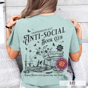 May include: A light teal t-shirt with a graphic design featuring a skull, books, and the text "ANTI-SOCIAL Book Club." The design includes butterflies, flowers, and the phrase "Making Friends With Characters, Not People."