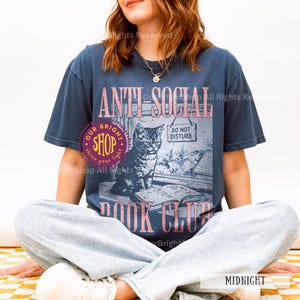 May include: Midnight blue t-shirt with a graphic featuring a cat, open book, and the words "ANTI-SOCIAL BOOK CLUB." The design includes a "DO NOT DISTURB" sign and the text "OUR BRIGHT SHOP."