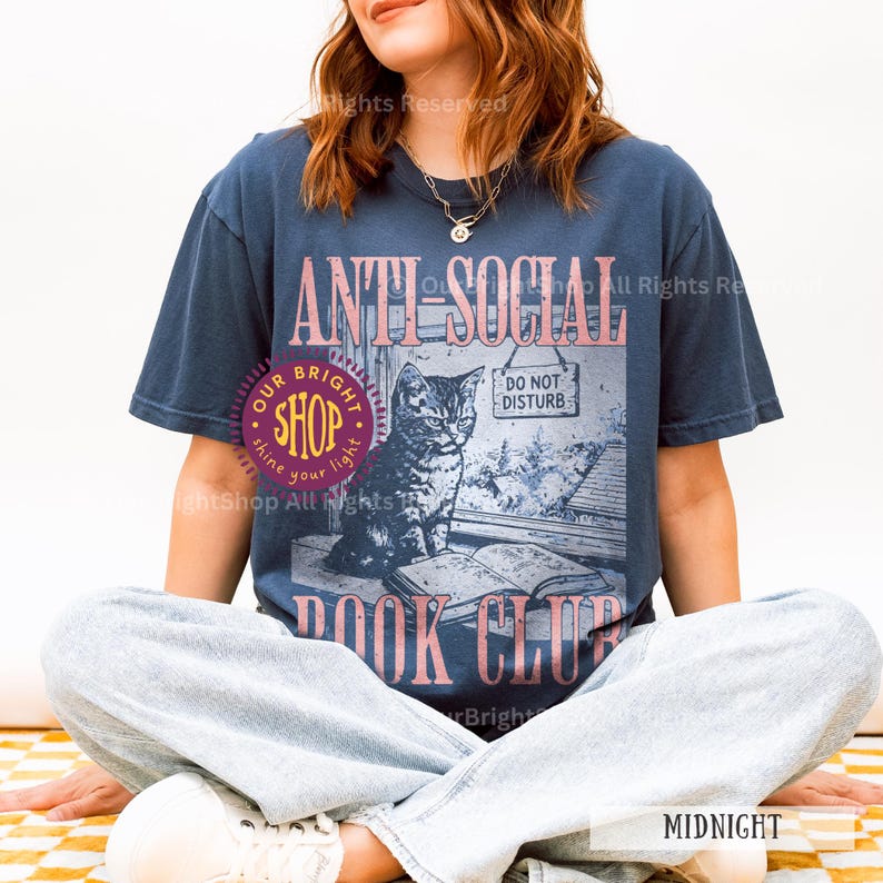 May include: Midnight blue t-shirt with a graphic featuring a cat, open book, and the words "ANTI-SOCIAL BOOK CLUB." The design includes a "DO NOT DISTURB" sign and the text "OUR BRIGHT SHOP."