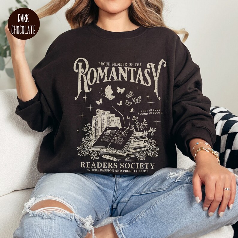 May include: A dark chocolate sweatshirt with a graphic design featuring the text "PROUD MEMBER OF THE ROMANTASY", a stack of books, butterflies, and the text "LOST IN LOVE FOUND IN BOOKS" and "READERS SOCIETY WHERE PASSION AND PROSE COLLIDE".