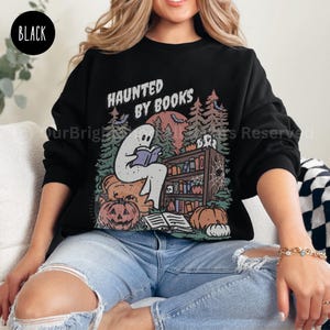 May include: Black sweatshirt with a Halloween-themed graphic. The design features a ghost reading a book in front of a bookshelf, pumpkins, and the text "HAUNTED BY BOOKS". The sweatshirt is a crew neck style.