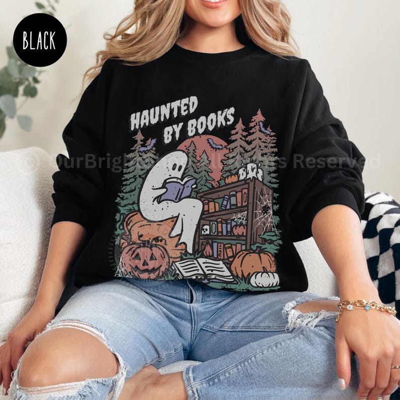 May include: Black sweatshirt with a Halloween-themed graphic. The design features a ghost reading a book in front of a bookshelf, pumpkins, and the text "HAUNTED BY BOOKS". The sweatshirt is a crew neck style.