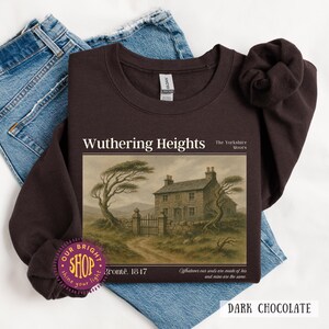 Puede incluir: Sudadera de color chocolate oscuro con el texto "Wuthering Heights" y una ilustraci&oacute;n de una casa y un paisaje. La sudadera tambi&eacute;n incluye el texto "The Yorkshire Moors", "Bront&euml;, 1847" y una cita. La sudadera se muestra sobre unos vaqueros azules.