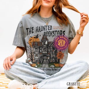 以下が含まれることがあります： お化け屋敷、カボチャ、そして「THE HAUNTED BOOKSTORS」の文字が描かれたグレーのTシャツ。デザインには、満月の前をほうきに乗って飛ぶ魔女が含まれています。シャツには「WITCH-OWN BOOKSTORS」の文字も入っています。