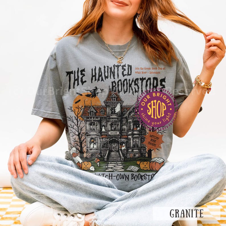 以下が含まれることがあります： お化け屋敷、カボチャ、そして「THE HAUNTED BOOKSTORS」の文字が描かれたグレーのTシャツ。デザインには、満月の前をほうきに乗って飛ぶ魔女が含まれています。シャツには「WITCH-OWN BOOKSTORS」の文字も入っています。