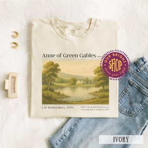 May include: Ivory-colored t-shirt featuring a landscape scene from "Anne of Green Gables." The design includes text and a quote. The shirt is paired with blue jeans, gold earrings, and a hair clip.