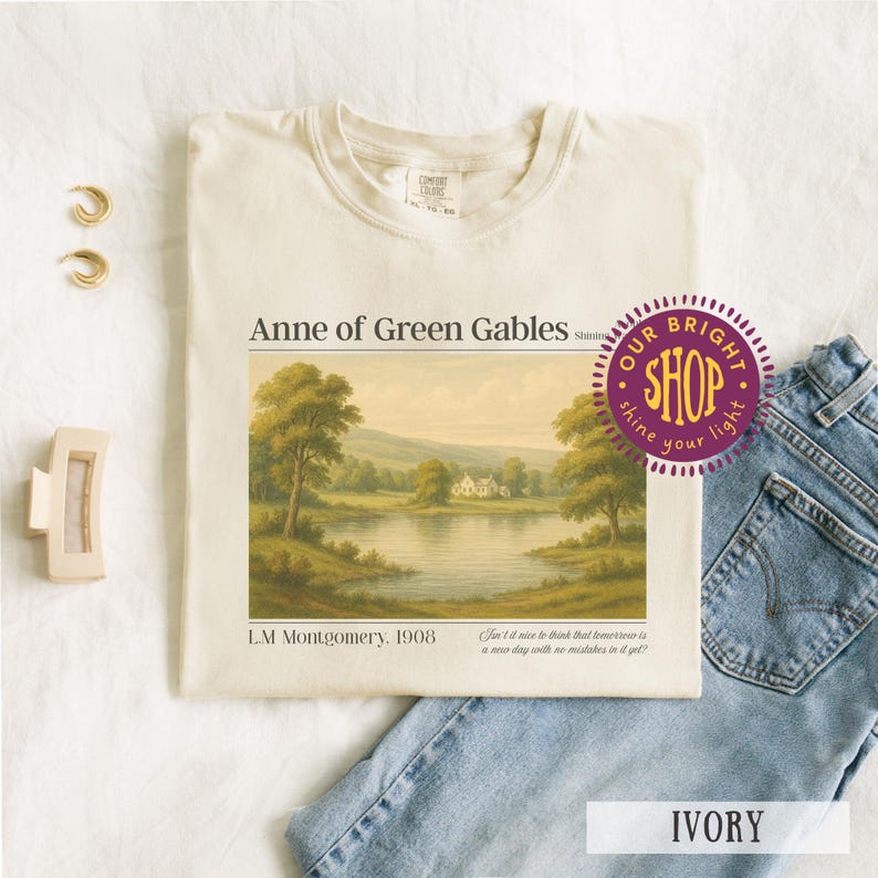 May include: Ivory-colored t-shirt featuring a landscape scene from "Anne of Green Gables." The design includes text and a quote. The shirt is paired with blue jeans, gold earrings, and a hair clip.