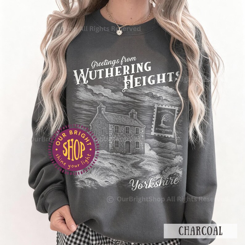 May include: Charcoal-colored sweatshirt featuring a graphic of a house and the text "Greetings from Wuthering Heights Yorkshire." The design includes a postage stamp illustration. The sweatshirt is a crew neck style.