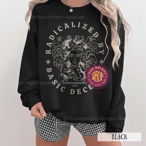 Peut inclure: Sweat-shirt noir avec un motif de M&eacute;duse entour&eacute;e de serpents, le texte "RADICALIZED BY BASIC DECENCY" dans un design circulaire et un logo de magasin violet. Le sweat-shirt porte &eacute;galement le texte "WOMEN'S RESISTANCE CLUB".