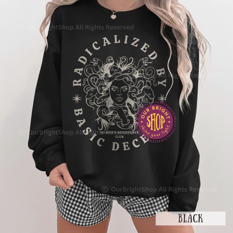 Peut inclure: Sweat-shirt noir avec un motif de M&eacute;duse entour&eacute;e de serpents, le texte "RADICALIZED BY BASIC DECENCY" dans un design circulaire et un logo de magasin violet. Le sweat-shirt porte &eacute;galement le texte "WOMEN'S RESISTANCE CLUB".