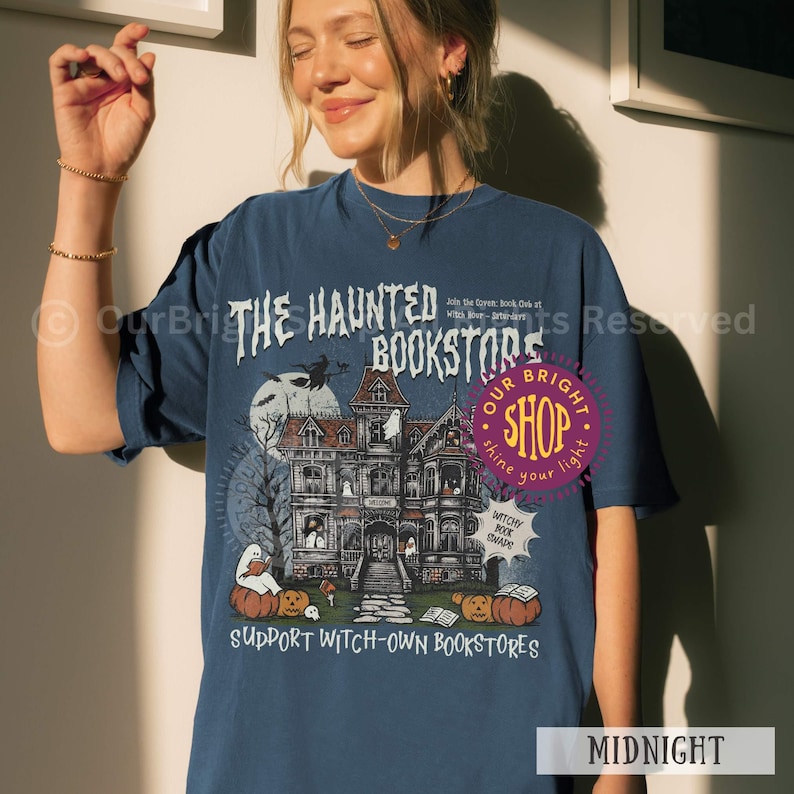 以下が含まれることがあります： お化け屋敷、カボチャ、そして「THE HAUNTED BOOKSTORE」の文字が描かれた、ハロウィーンをテーマにしたネイビーブルーのTシャツ。デザインには満月と魔女が含まれています。シャツには「SUPPORT WITCH-OWN BOOKSTORES」とも書かれています。