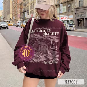 May include: Maroon sweatshirt with a graphic design featuring the text "Greetings from Wuthering Heights Yorkshire". The design includes an illustration of a house and a postage stamp. The sweatshirt is a solid maroon color.