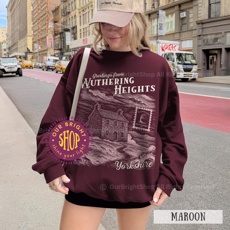 May include: Maroon sweatshirt with a graphic design featuring the text "Greetings from Wuthering Heights Yorkshire". The design includes an illustration of a house and a postage stamp. The sweatshirt is a solid maroon color.