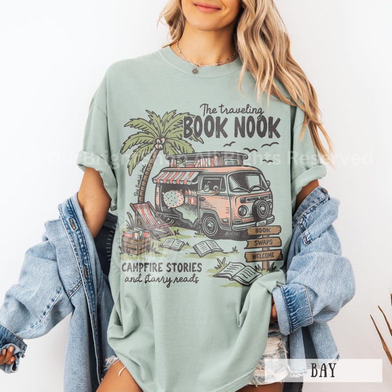 May include: A teal blue t-shirt with a graphic of a vintage van with a sign that says "The Traveling Book Nook". The van is decorated with books and has a palm tree next to it. The text "Campfire Stories and Starry Reads" is printed below the van.