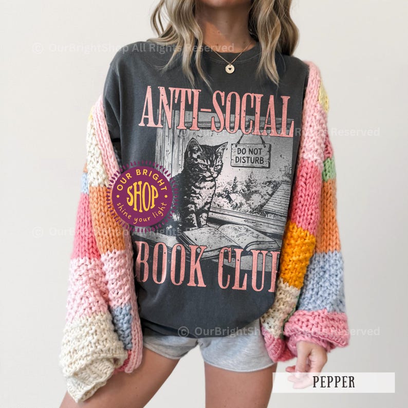 May include: Dark gray t-shirt with the text "ANTI-SOCIAL BOOK CLUB" and a graphic of a cat by a window. The person is wearing a colorful, striped cardigan with pink, yellow, and blue blocks.