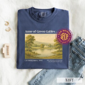 May include: Navy blue t-shirt featuring a landscape scene from "Anne of Green Gables." The graphic includes text: "Anne of Green Gables," "L.M. Montgomery, 1908," and a quote. A "Our Bright Shop" sticker is visible. The shirt is paired with blue jeans.