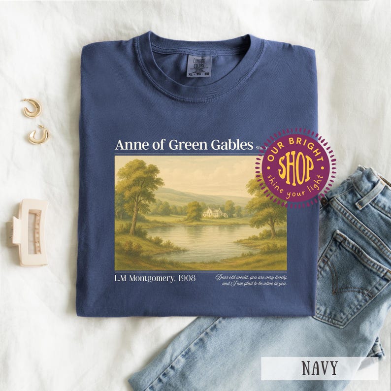 May include: Navy blue t-shirt featuring a landscape scene from "Anne of Green Gables." The graphic includes text: "Anne of Green Gables," "L.M. Montgomery, 1908," and a quote. A "Our Bright Shop" sticker is visible. The shirt is paired with blue jeans.