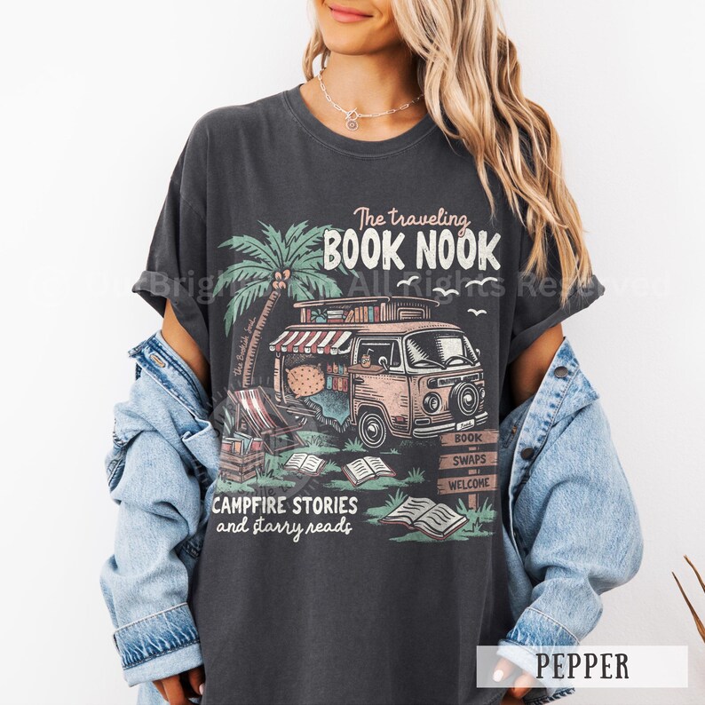 May include: A dark gray t-shirt with a graphic print of a vintage van with a sign that reads "The Traveling Book Nook". The van is filled with books and has a sign that says "Campfire Stories and Starry Reads".