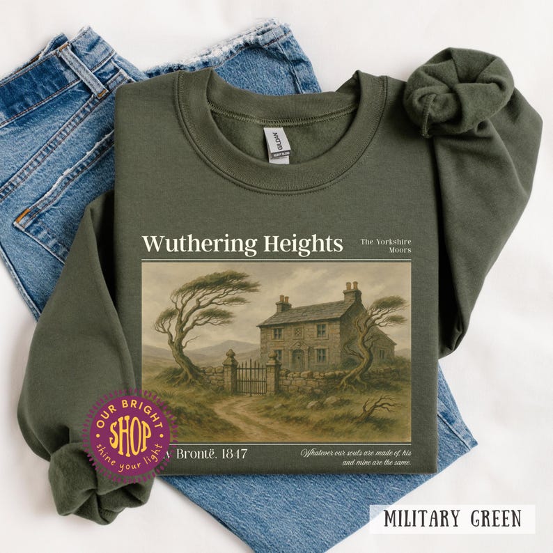 Puede incluir: Sudadera verde militar con el texto "Wuthering Heights" y una ilustraci&oacute;n de un paisaje. La sudadera tambi&eacute;n incluye el texto "The Yorkshire Moors", "Bront&euml;. 1847" y una cita. La sudadera se muestra sobre unos vaqueros azules.