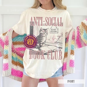 May include: Ivory-colored t-shirt with the text "ANTI-SOCIAL BOOK CLUB" and a graphic of a cat sitting on a book. The shirt also features a "DO NOT DISTURB" sign and a shop logo. The model is wearing a colorful, striped cardigan.
