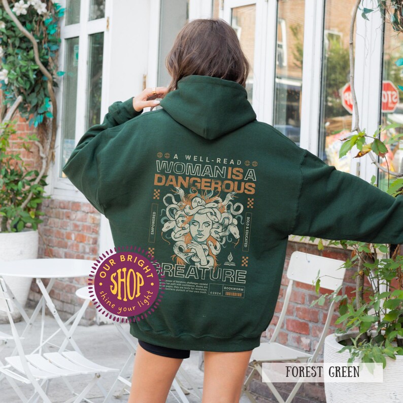 Op de afbeelding: Donkergroene hoodie met de tekst "A well-read woman is a dangerous creature" en een Medusa-illustratie. Het ontwerp is in beige en groene tinten. De hoodie heeft een capuchon en lange mouwen.