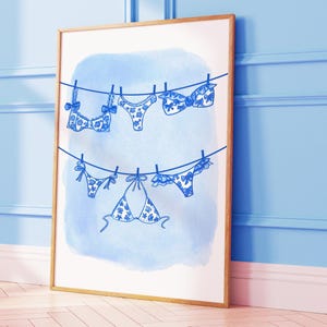 May include: A framed art print featuring a blue watercolor background with a line of blue and white patterned lingerie hanging on a clothesline. The lingerie includes bras and bikini bottoms, with bow details.
