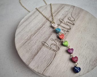 Könnte beinhalten: Eine goldene Kettenkette mit einer Reihe von bunten herzförmigen Steinen. Die Steine sind in den Farben Rosa, Blau, Orange, Grün, Lila und Rot. Die Kette wird auf einer Holzoberfläche mit dem Wort "atmen" darauf präsentiert.