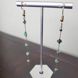 May include: A pair of long gold chain earrings with turquoise stones. The earrings are hanging from a silver stand.