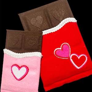 May include: Two felt chocolate bar-shaped decorations. One is pink with a white and red heart, the other is red with two hearts. The top portion is brown with heart cutouts, and the bottom has white trim.