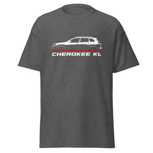 May include: Dark grey t-shirt with a white side profile of a Jeep Cherokee KL SUV. The design includes a red stripe and the text "CHEROKEE KL" in white, centered below the vehicle graphic. The shirt is made of a soft material.