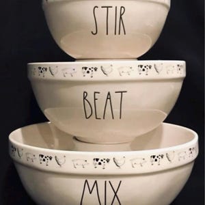 May include: A set of three white ceramic mixing bowls with black cow and chicken designs around the rim. The bowls are labeled "Mix", "Beat", and "Stir".