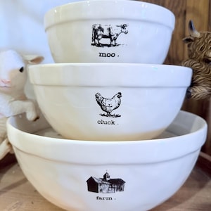 May include: Three white ceramic bowls stacked on top of each other. Each bowl has a black graphic and text: a cow with "moo", a chicken with "cluck", and a barn with "farm".