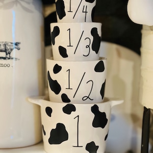 May include: Set of four white ceramic measuring cups with black cow print design. The cups are stacked in order from largest to smallest, with the following fractions printed on them: 1, 1/2, 1/3, and 1/4.