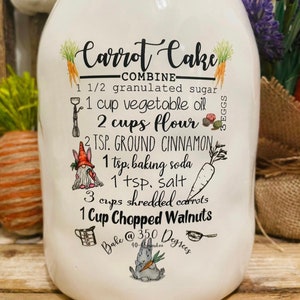 May include: White ceramic jar with a carrot cake recipe printed on it. The recipe includes ingredients like sugar, flour, cinnamon, and walnuts. Decorative illustrations of carrots, a gnome, and a bunny are included.