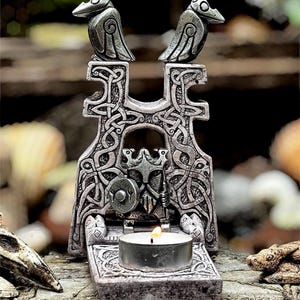 Könnte beinhalten: Ein detaillierter, grau getönter Kerzenhalter mit zwei Vogelfiguren, die auf einem keltischen Knotenmuster sitzen. Der Kerzenhalter weist aufwendige Schnitzereien und eine brennende Teelichtkerze auf.
