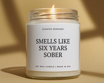 Può includere: Una candela di cera di soia in un barattolo di vetro trasparente con uno stoppino acceso. L'etichetta bianca riporta il testo Scented Serenade, Smells Like Six Years Sober e Soy Wax Candle | Made in USA in nero. La cera è color bianco sporco.