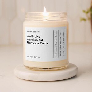 May include: A lit soy wax candle in a clear glass jar with a white label. The text reads: Scented Serenade, Smells Like World's Best Pharmacy Tech, Hand-poured Soy Wax Candle, 50-60 Hours Burning Time, Eco-Friendly & Non-Toxic, KEEP THAT SHIT UP.
