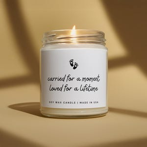 Op de afbeelding: Een sojawas kaars in een doorzichtige glazen pot met een wit etiket met twee kleine voetafdrukken met hartjes en de tekst: carried for a moment loved for a lifetime. De kaars brandt en toont een kleine vlam aan de bovenkant.