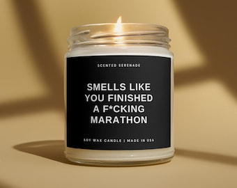 May include: A clear glass jar candle with a black label featuring white text that reads: SCENTED SERENADE, SMELLS LIKE YOU FINISHED A F*CKING MARATHON, SOY WAX CANDLE | MADE IN USA. The candle has a lit wick and contains white soy wax.