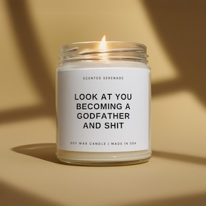 May include: A lit soy wax candle in a glass jar. The candle has a white label with the text "SCENTED SERENADE" and "LOOK AT YOU BECOMING A GODFATHER AND SHIT". The candle is made in the USA.