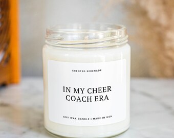 May include: A clear glass jar candle filled with white soy wax. The white rectangular label features the text SCENTED SERENADE at the top, IN MY CHEER COACH ERA in the center, and SOY WAX CANDLE | MADE IN USA at the bottom.