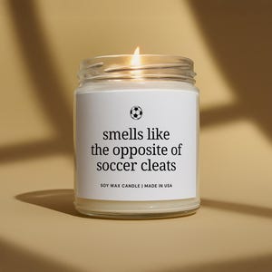 May include: A clear glass jar soy wax candle with a white label featuring a black soccer ball icon and the text: smells like the opposite of soccer cleats. Below the text, it reads: SOY WAX CANDLE | MADE IN USA. The candle has a lit wick.