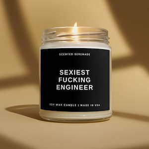 May include: A clear glass jar candle with a lit wick and a black label. The label features the white text SCENTED SERENADE, SEXIEST FUCKING ENGINEER, and SOY WAX CANDLE | MADE IN USA. The candle contains off-white wax and is in a cylindrical glass container.