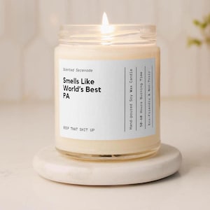 May include: A clear glass jar candle with a white label featuring the text: Scented Serenade, Smells Like World's Best PA, KEEP THAT SHIT UP, Hand-poured Soy Wax Candle, 50-60 Hours Burning Time, and Eco-Friendly & Non-Toxic. The candle has cream wax.